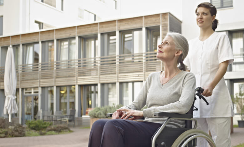 Alte Dame im Rollstuhl, geschoben von einer Pflegekraft vor einem Seniorenheim